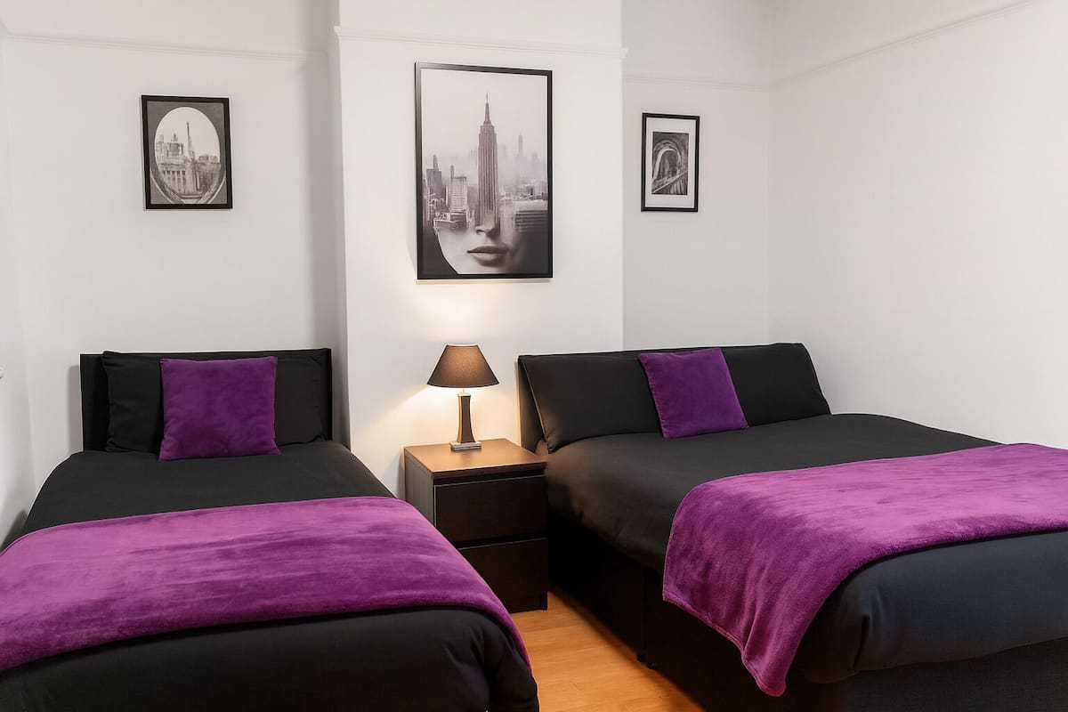 A hotel room with two beds, each with black and purple bedding, a nightstand with a lamp, and framed cityscape pictures on the walls.