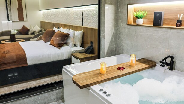 A modern bedroom with a large bed, bedside tables, a nightstand, a wooden shelf with a plant, and a bathtub with candles. - Royal Regency Hotel EG (New Cairo)