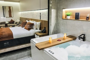 A modern bedroom with a large bed, bedside tables, a nightstand, a wooden shelf with a plant, and a bathtub with candles.