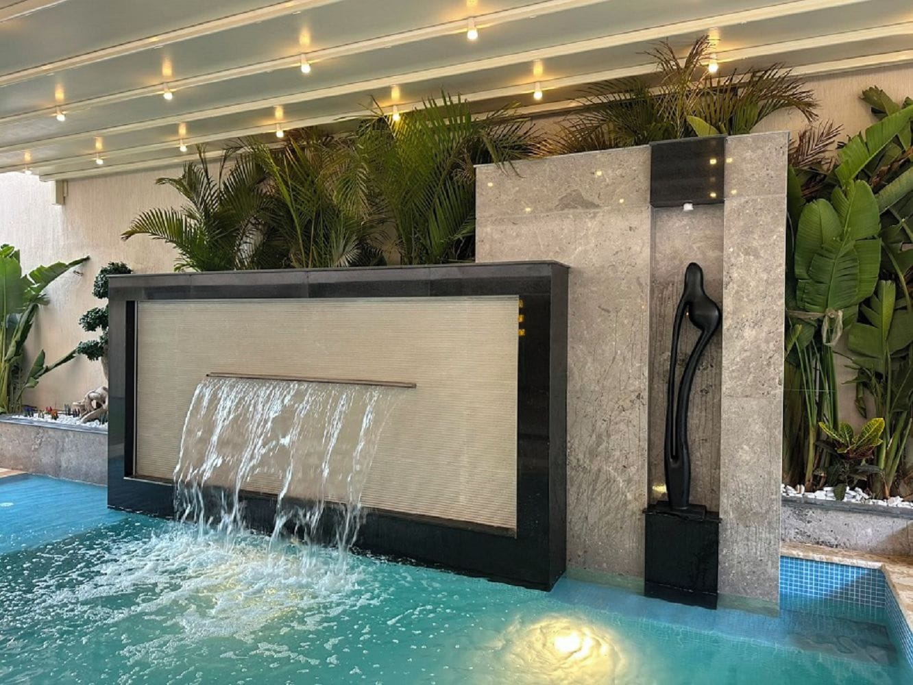 A pool area with a waterfall feature, surrounded by tropical plants and a stone wall.