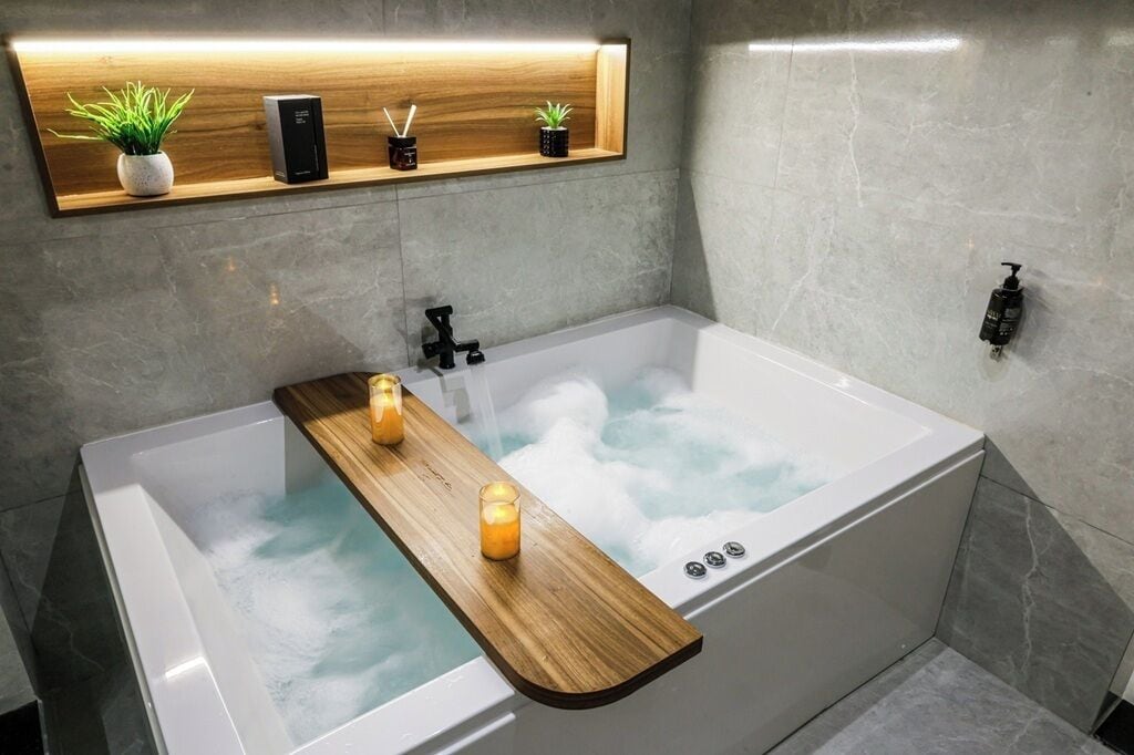 A modern bathroom with a large bathtub, a wooden shelf with plants and toiletries, and a handheld showerhead.