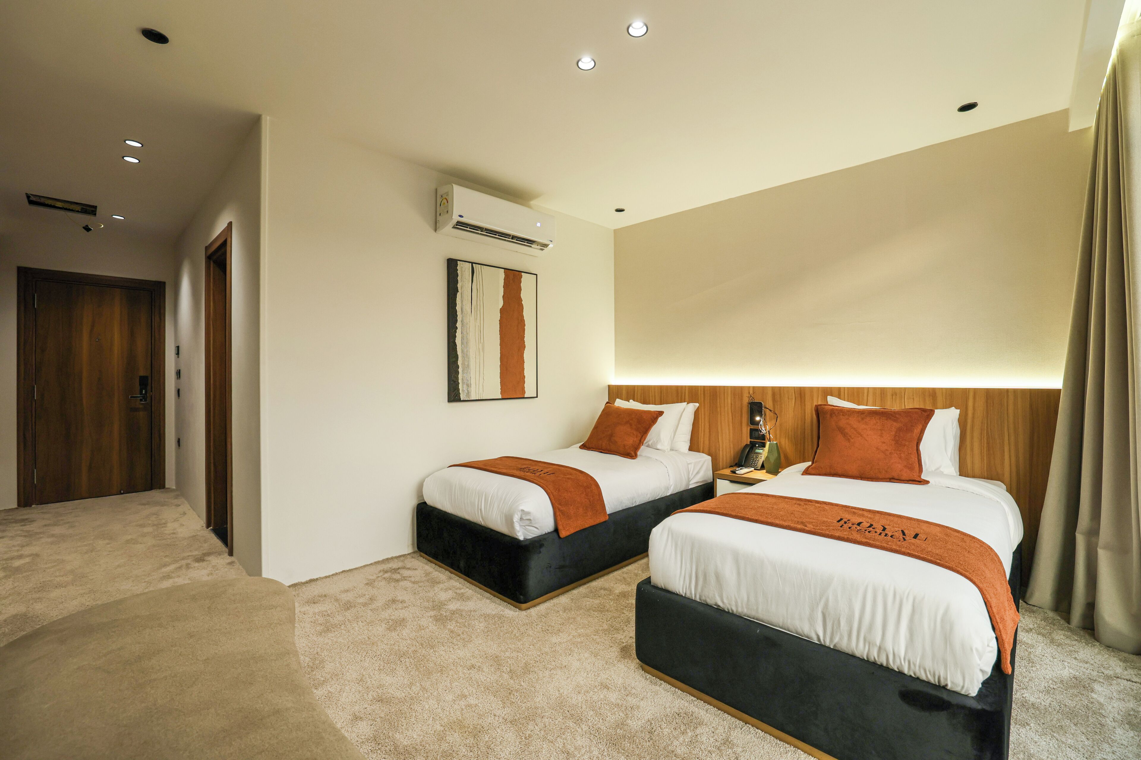 A hotel room with two beds, a wooden door, an air conditioning unit, and a painting on the wall.
