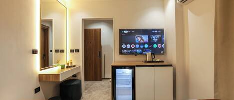 Habitación de hotel con cama, mesita de noche, espejo, televisor, refrigerador pequeño y puerta.