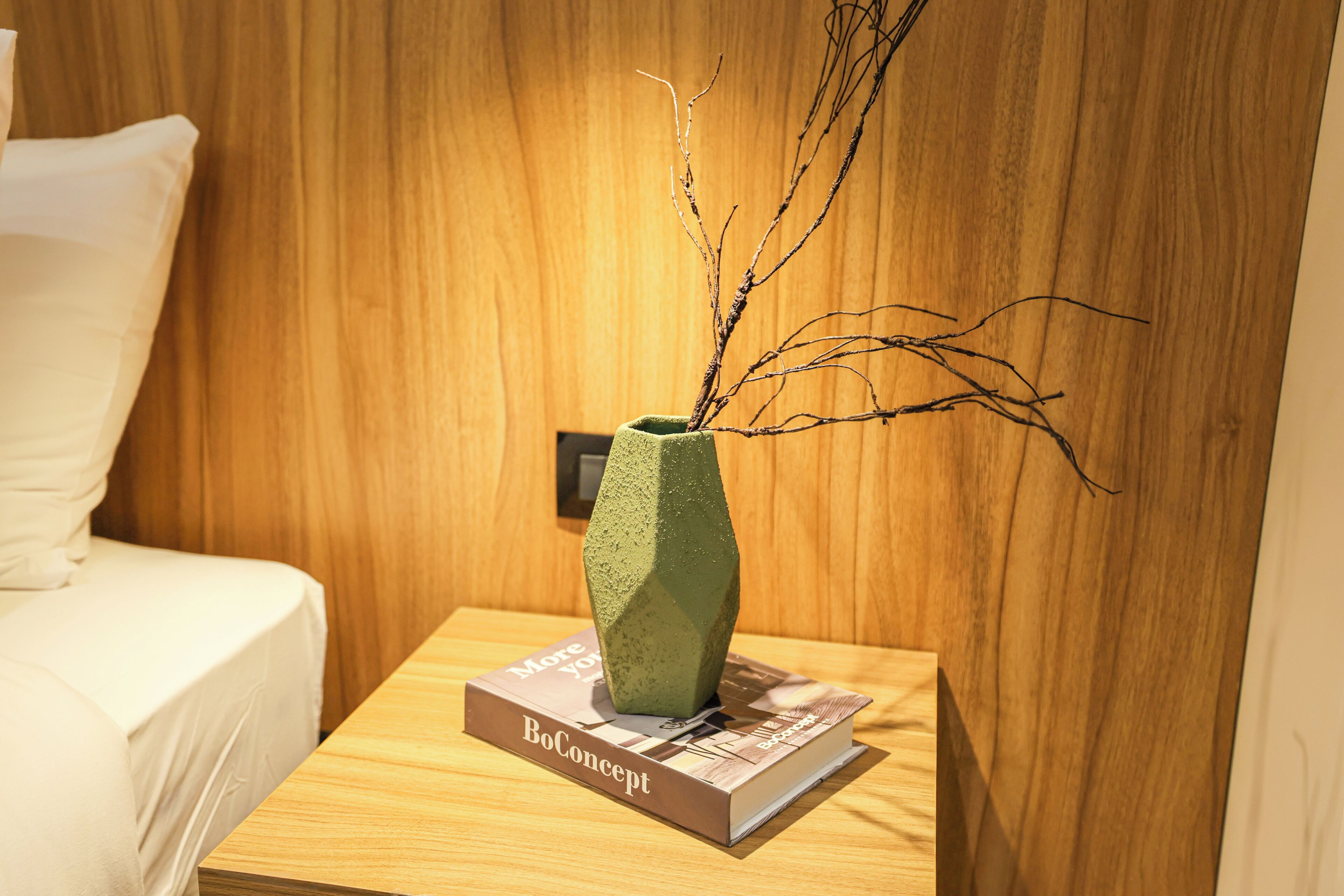 Ein Nachttisch aus Holz mit einem grünen Strauß in einer Vase, einem Buch mit dem Titel 'BoConcept' und einem weißen Kopfkissen auf einem Bett.