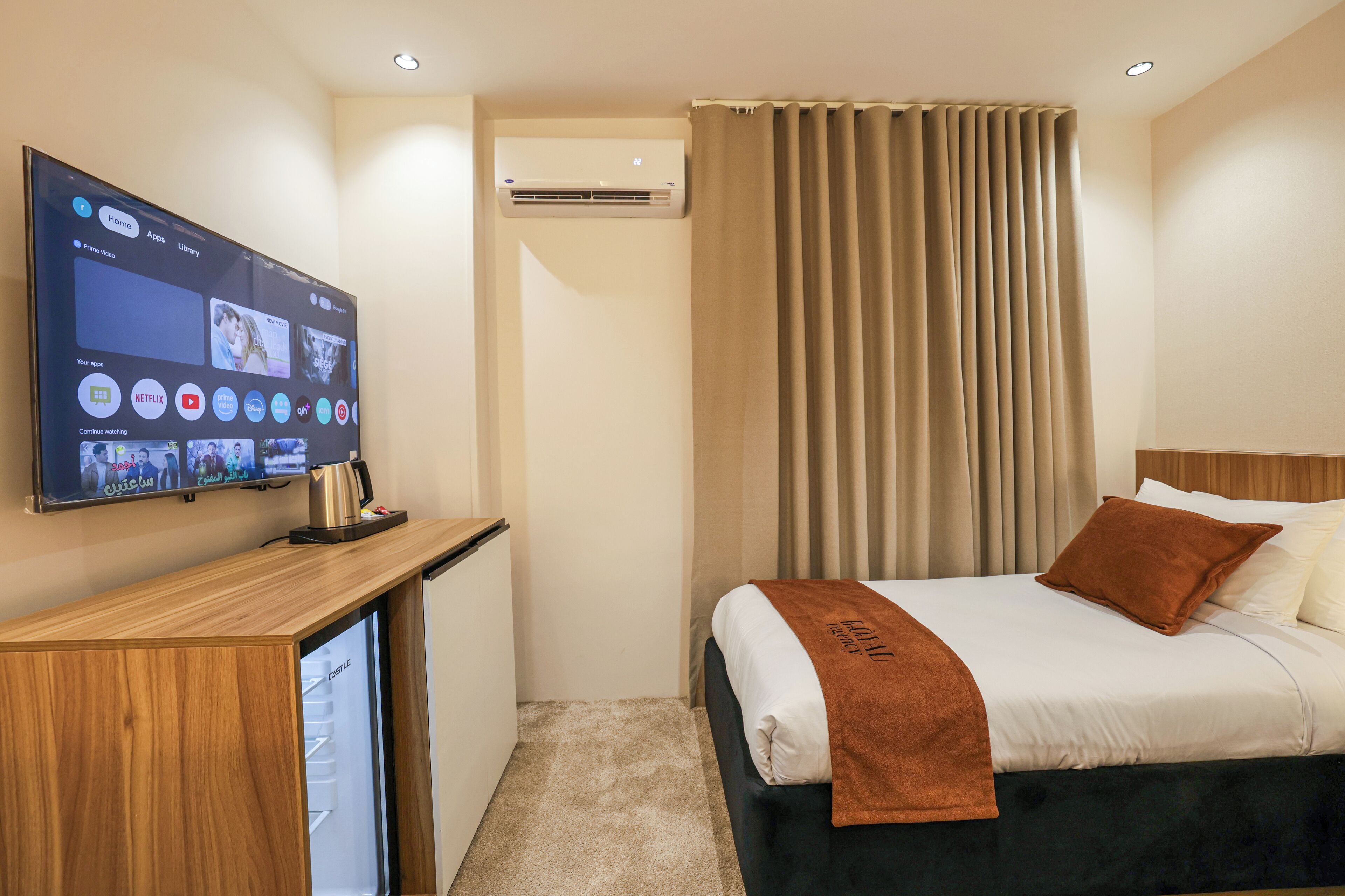 A hotel room with a bed, a TV mounted on the wall, a desk with a coffee machine, and a window with curtains.