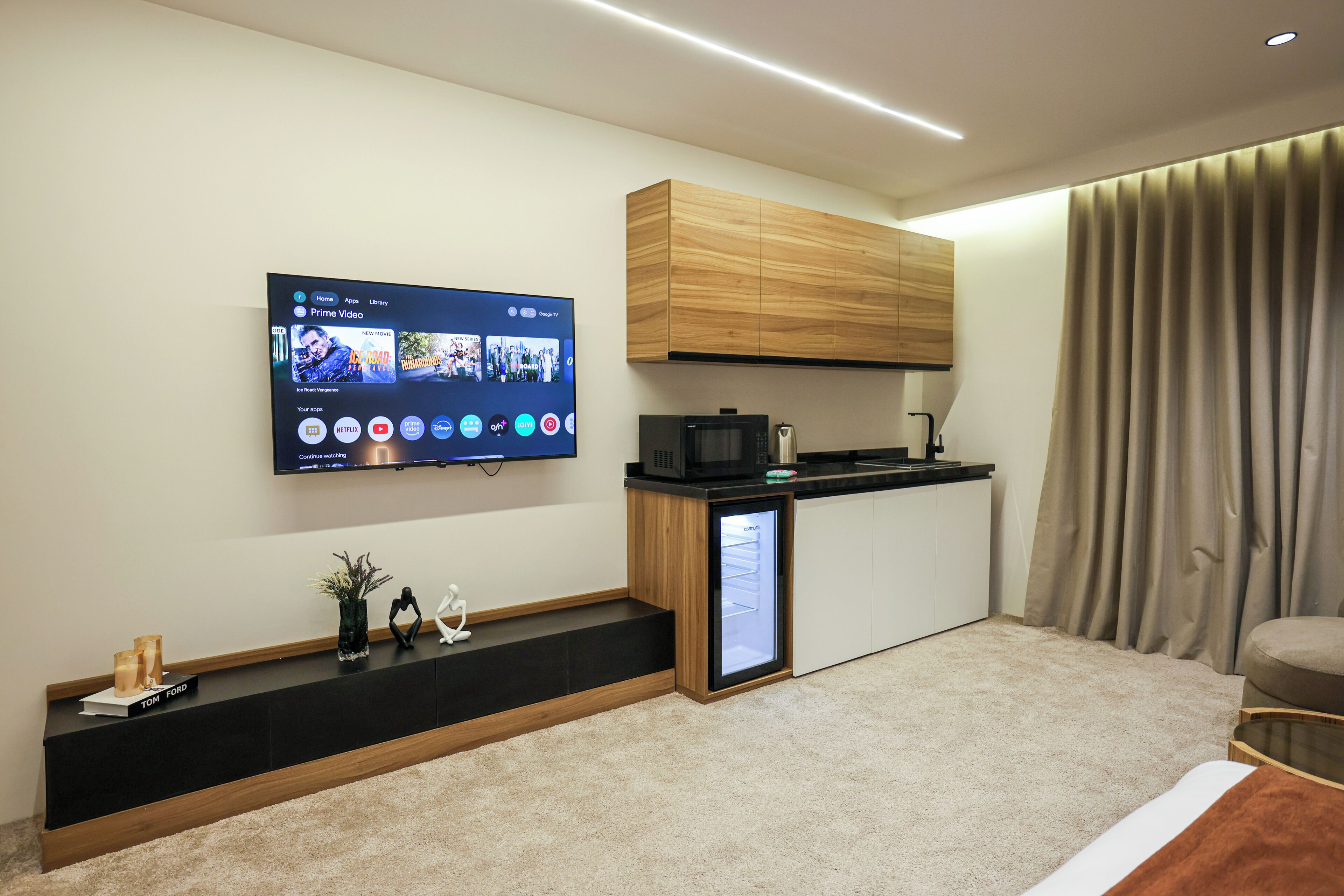 En moderne opholdsstue med et fladskærms TV på væggen, et integreret tekøkken med mikroovn, samt et siddeområde med sofa og sofabord.