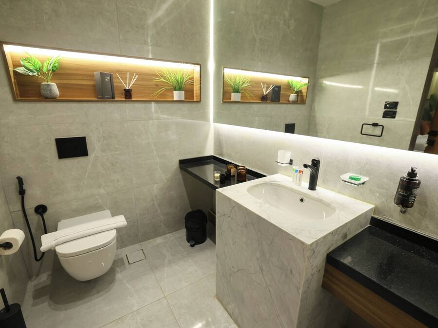 Un baño moderno con un inodoro blanco, un lavabo con encimera de mármol y dos estantes empotrados con plantas y artículos de aseo.