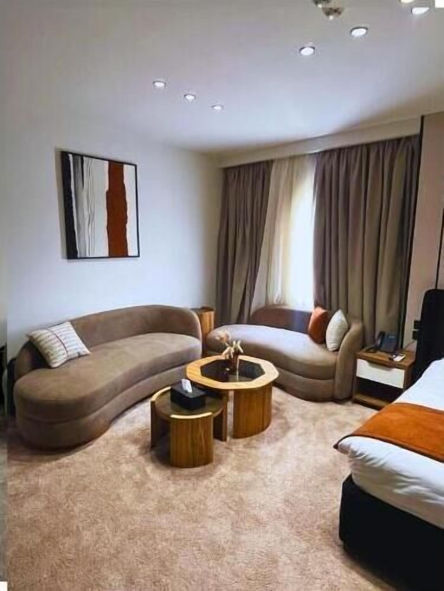 A modern hotel room with a curved sofa, a round coffee table, a bed, and a large window with curtains.