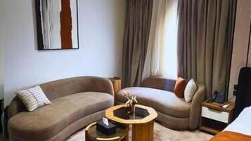 Une chambre d’hôtel moderne avec un canapé incurvé, une table basse ronde, un lit et une grande fenêtre avec des rideaux.