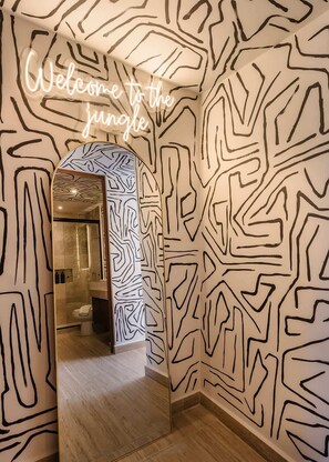 A room with a unique wall pattern, a neon sign reading 'Welcome to the jungle,' and a mirror reflecting a bathroom with a modern design.