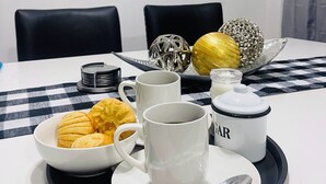 Una mesa de comedor con mantel a cuadros, dos sillas negras, un plato blanco con una cuchara, un bol de pasteles, dos tazas blancas, una jarrera blanca etiquetada “GAR” y objetos decorativos sobre una bandeja.