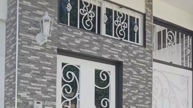 Fachada de edificio con diseño moderno, que presenta un muro de piedra texturizada, detalles de metal decorativo alrededor de las ventanas y la puerta, y dos grandes macetas con plantas.