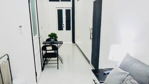 Un pasillo estrecho con una cama al fondo, una mesita con una silla y una puerta que da a otra habitación.