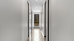 A modern hallway with white walls and a polished floor, leading to a large window with a view of trees.