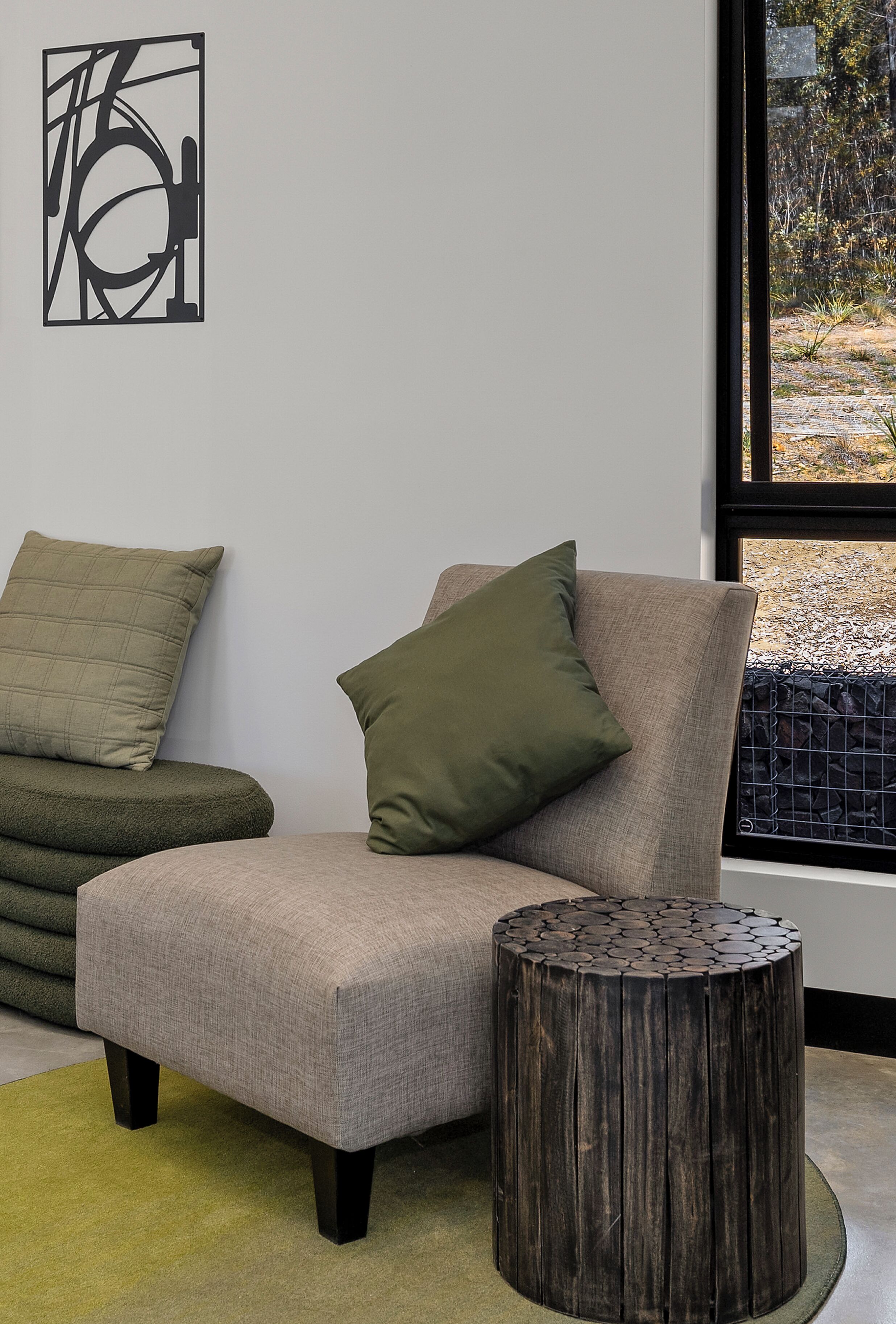 A modern living room with a grey sofa, a green pillow, a round wooden side table, and a large window with a view of a garden.