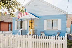 Una casa azul con cerca blanca de piquete, una puerta amarilla y dos sillas afuera.