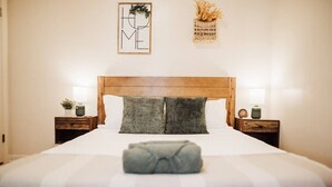 Un llit ben fet amb un edredó estriat, dos coixins i una manta doblegada al capdamunt. Hi ha dues taules de nit amb làmpades i objectes decoratius. Una obra d'art emmarcada amb la paraula “HOME” hi ha a la paret sobre el llit.