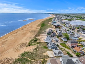 Une vue aérienne d’une zone résidentielle côtière, avec des maisons le long de la plage, un ciel dégagé et une mer calme.