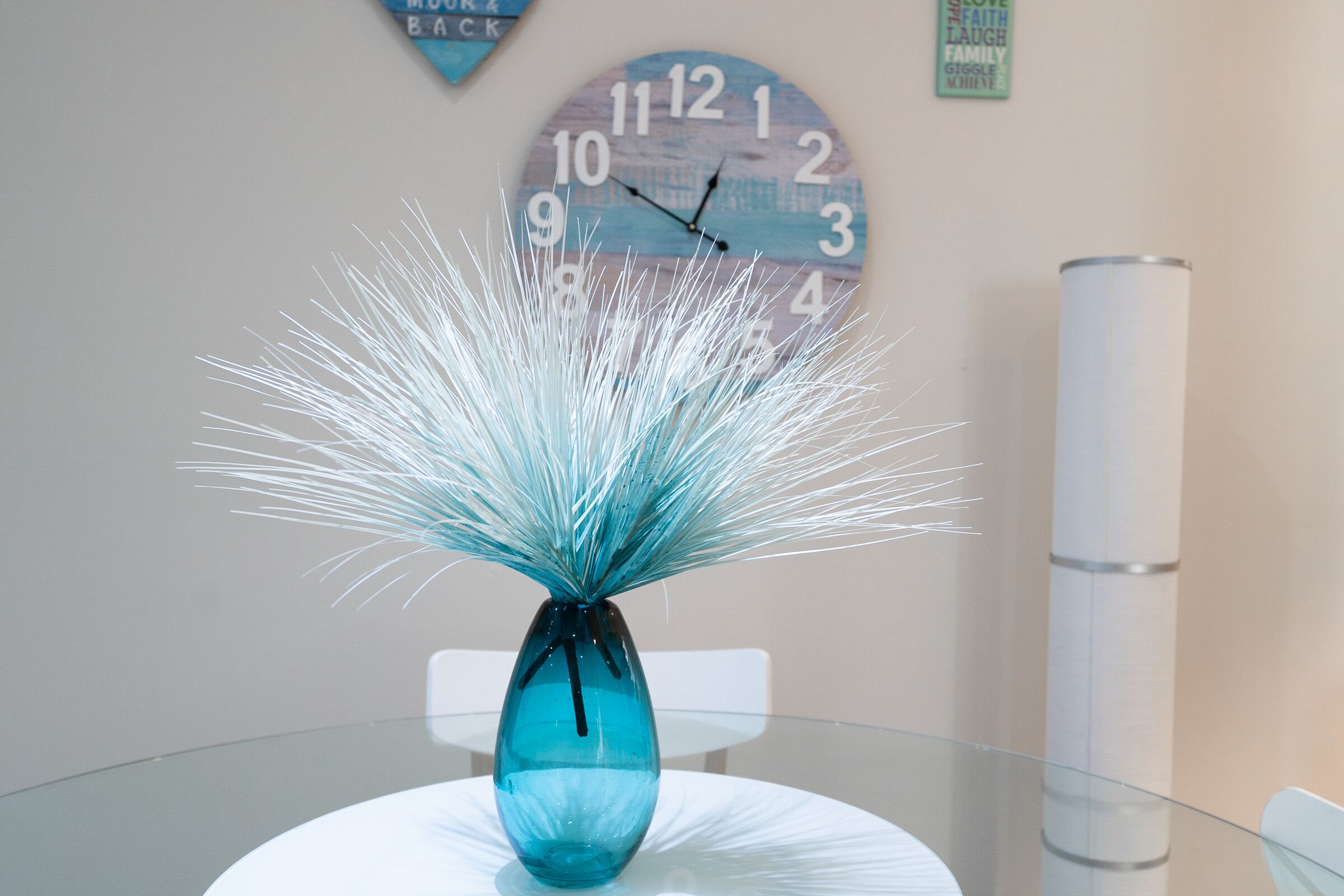 A glass table with a blue vase containing white decorative elements, a clock with a blue and white design, and a white chair.