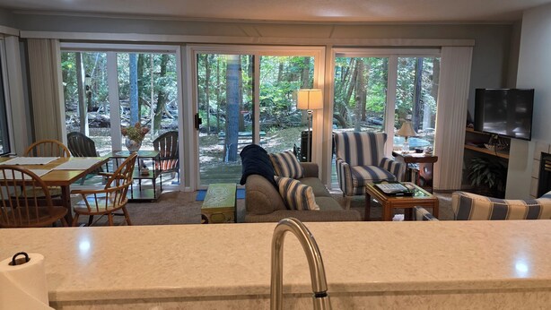A modern living room with a large sliding glass door leading to a deck, a sofa, a coffee table, and a TV mounted on the wall. - Creek Comfort! Charming sleeps 6 condo in lovely Bellaire. Short drive to skiing (Bellaire)