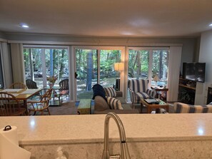 Ein modernes Wohnzimmer mit einem großen Schiebe-Glasfenster, das auf eine Terrasse führt, einer Couch, einem Couchtisch und einem an der Wand befestigten Fernseher.