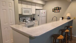 A modern kitchen with white cabinets, a built-in stove, a microwave, and a refrigerator. There are three wooden stools at the bar counter. - Creek Comfort! Charming sleeps 6 condo in lovely Bellaire. Short drive to skiing (Bellaire)