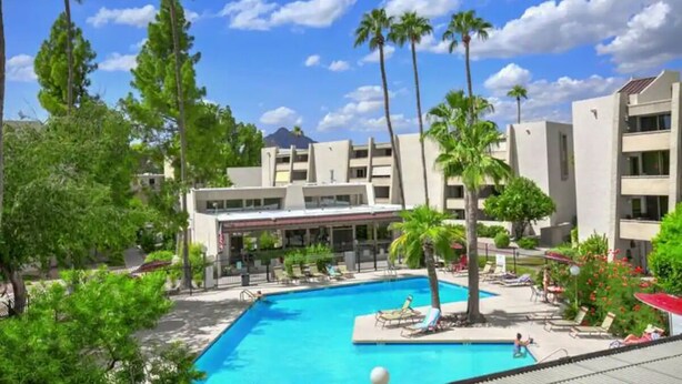 Zona de piscina con palmeras, sillas de descanso y un edificio de varios pisos al fondo.