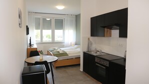 Et lille hotelværelse med seng, et bord med vase, tekøkken med komfur og vask samt et vindue med gardiner.