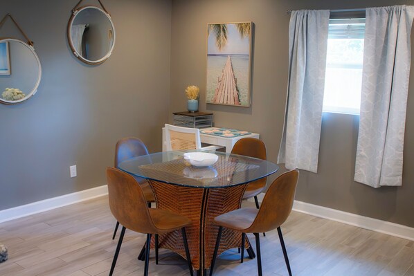 Un comedor con mesa de vidrio rodeada de sillas de mimbre, un cuadro en la pared y una ventana con cortinas.