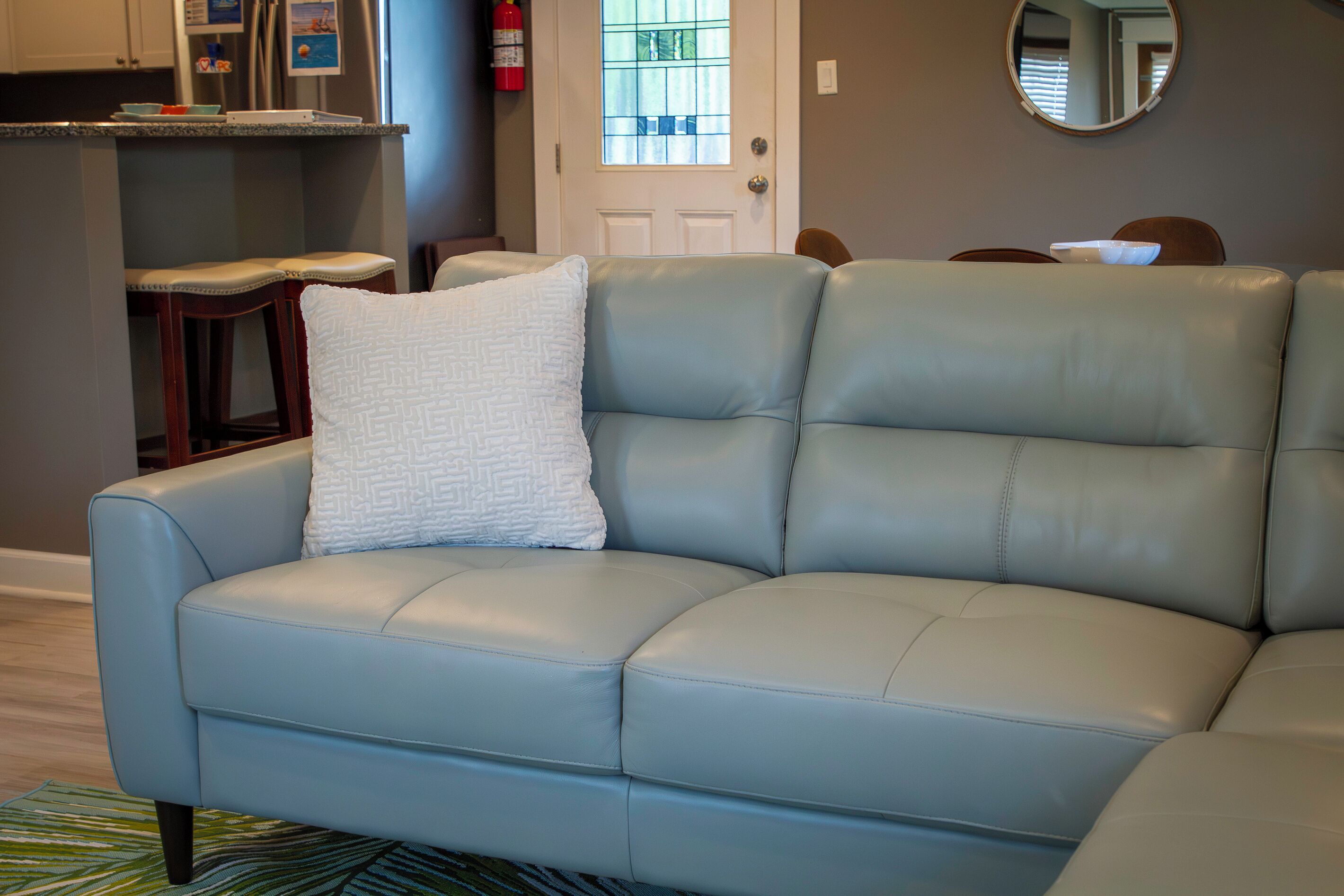 A light blue leather sofa with a white cushion, a wooden chair, a round mirror, and a kitchen area with a refrigerator and a fire extinguisher.