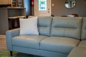 A light blue leather sofa with a white cushion, a wooden chair, a round mirror, and a kitchen area with a refrigerator and a fire extinguisher.
