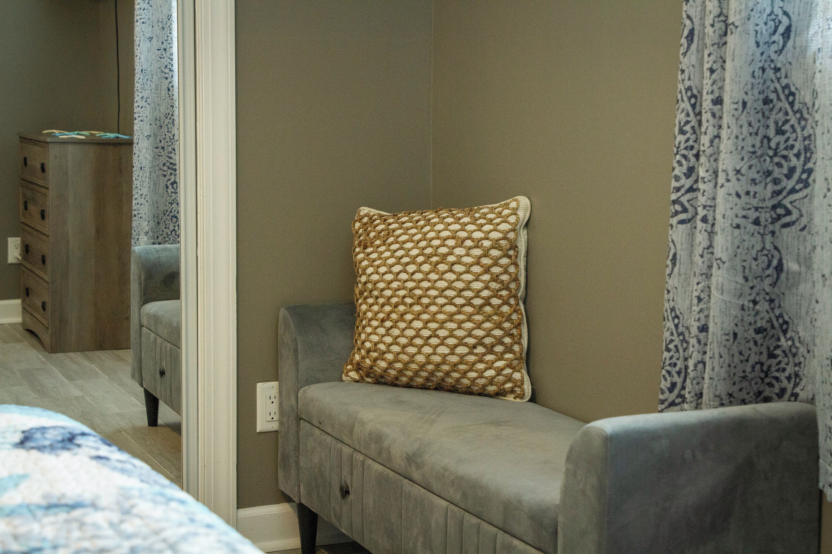 Un dormitorio con un sofá gris, un cojín estampado, una mesita de noche y un espejo.