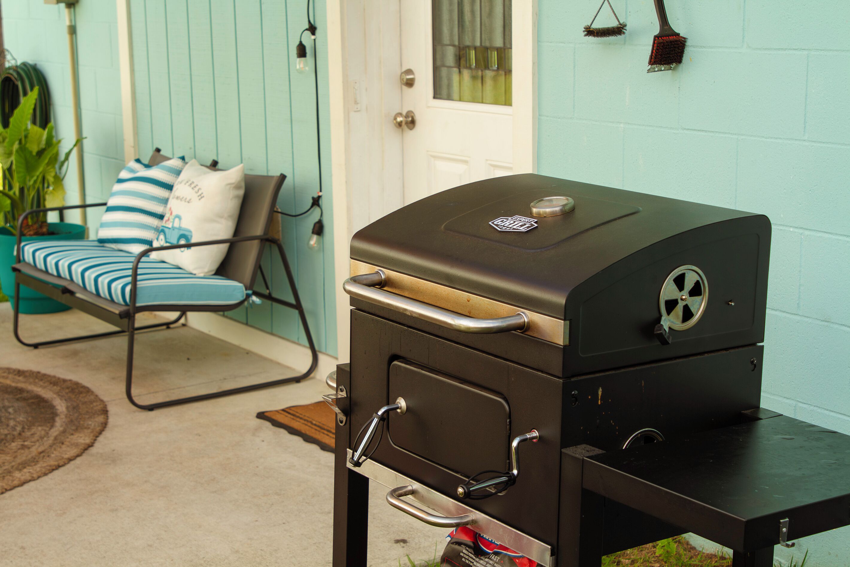 A barbecue grill with a handle and a wheel lock, placed on a patio with a chair and cushions.