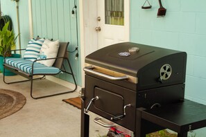 Una parrilla para barbacoa con asa y un seguro con ruedas, ubicada en un patio con una silla y cojines.