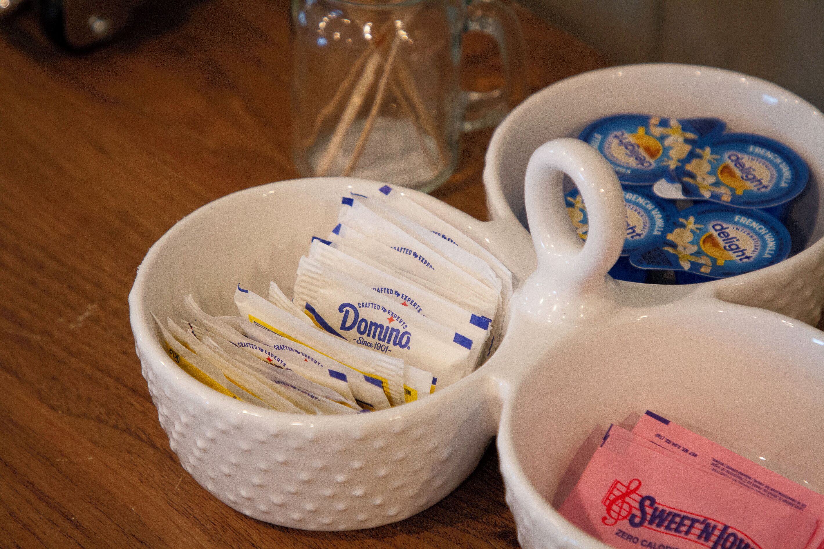 A white ceramic mug with a handle, filled with white packets labeled 'Domino' and a pink packet labeled 'Sweet'N Low'.