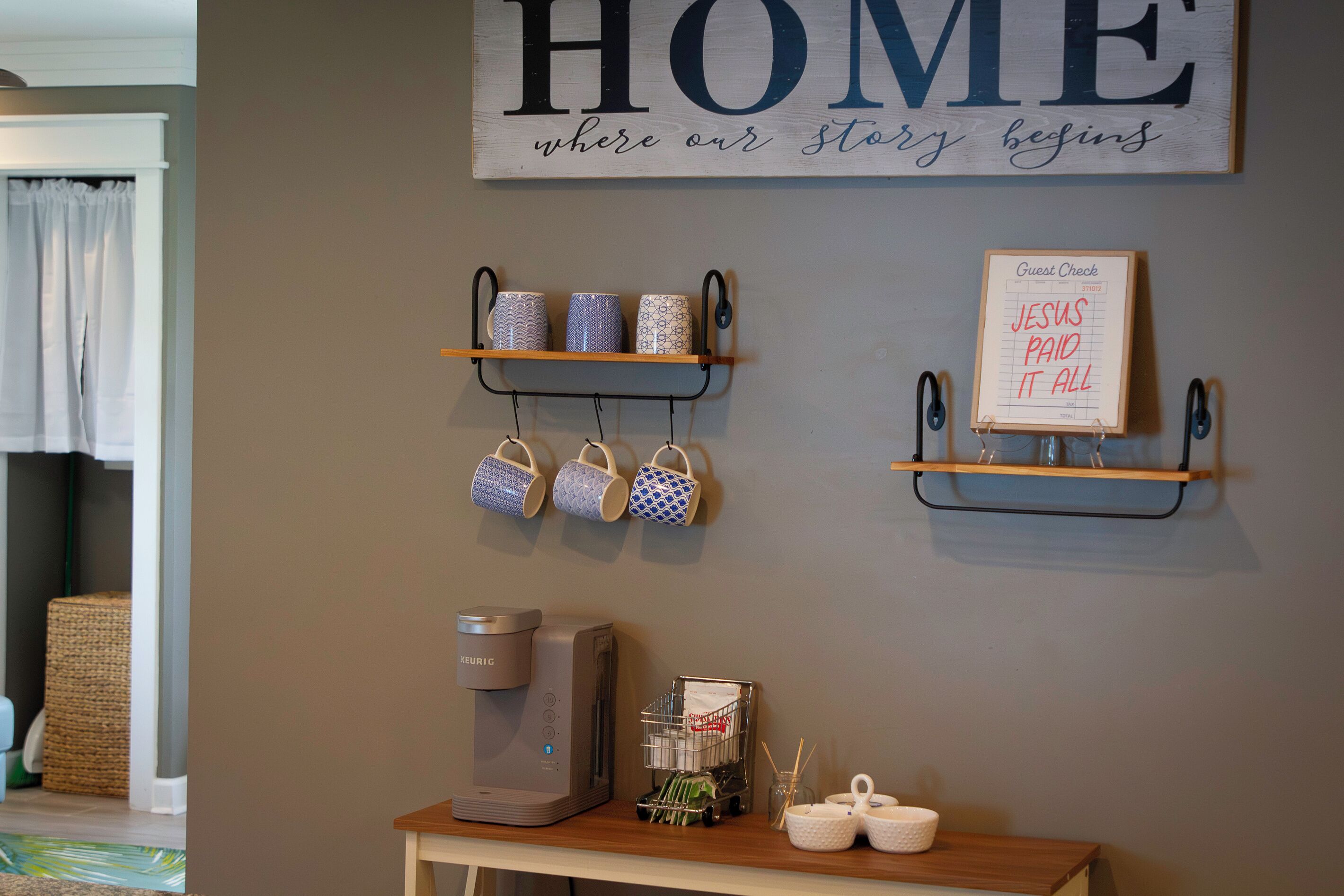 A wall with a sign that reads 'HOME where our story begins' and a 'Guest Check' board with 'JESUS PAID IT ALL' written on it.