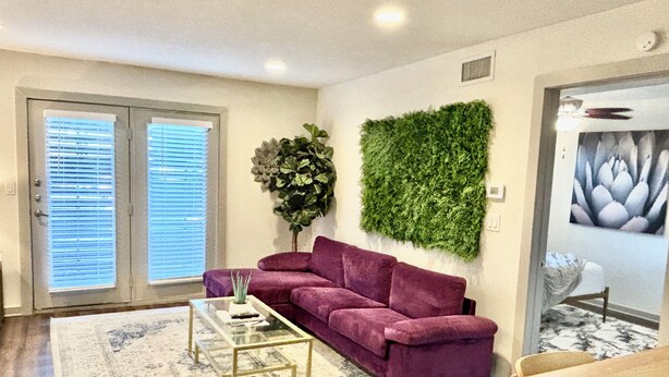 A living room with a purple sofa, a glass coffee table, a green wall hanging, and a dining area with a wooden table.