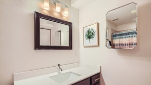 A bathroom with a white sink, dark cabinets, a large mirror, and a framed plant.
