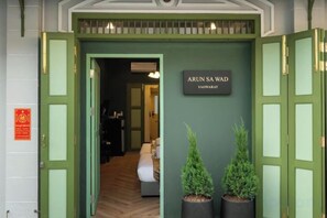 L’entrée d’un hôtel, avec une porte verte, une enseigne indiquant « ARUN SA MAD VAORAWAT » et deux plantes en pot.
