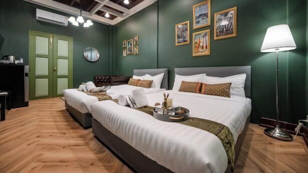 Habitación de hotel con dos camas, pared verde, cuadros enmarcados, lámpara y suelo de madera.