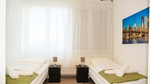 Ett hotellrum med två separata sängar, ett stort fönster med tunna, genomskinliga gardiner, en tavla som föreställer en stadsbild och en grön pläd på varje säng.