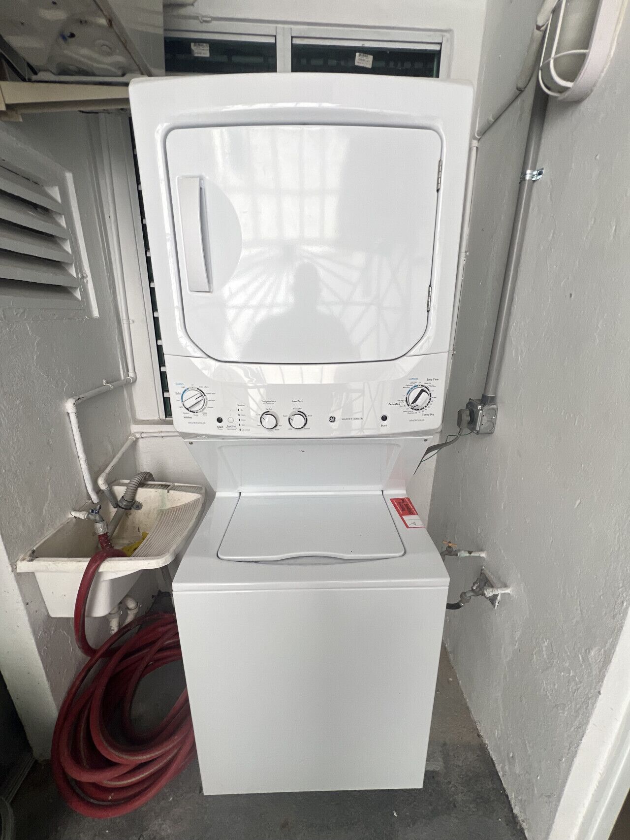 A compact, white washing machine with a control panel, a red hose, and a small sink.