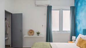 Une chambre à coucher comprenant un lit, une table de chevet, un climatiseur fixé au mur, une fenêtre avec des rideaux, une étagère avec un chapeau et une plante, et une porte donnant sur un placard.