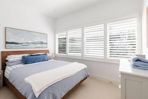 A bedroom with a bed, a window with blinds, a painting on the wall, and a bedside table.