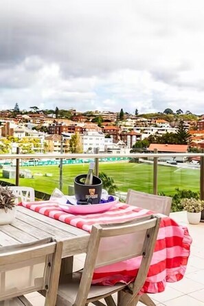 A balcony with a table and chairs, a potted plant, and a view of a green field and buildings in the background.