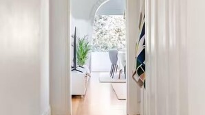 A hallway with wooden flooring, white walls, and a door leading to a room with a white sofa and a plant. - Dolphin Sands; A 3-bedroom apartment in Coogee Beach, barefoot walk to sand. (Coogee)