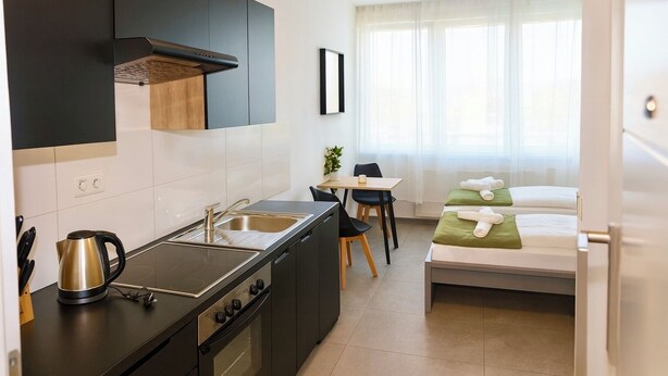 Cocina compacta con estufa, pileta y tetera, una pequeña mesa de comedor con sillas, y una cama con ropa de cama verde.