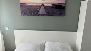 Una cama con ropa de cama blanca, una pared gris y una pintura de atardecer encima.