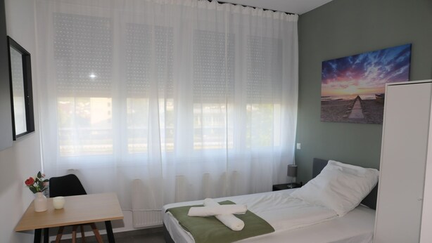 Una habitación de hotel con una cama, una mesa, una silla, un cuadro y una ventana con cortinas traslúcidas.