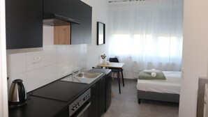 Cocina compacta con estufa, pileta y muebles negros. Hay un pequeño comedor con mesa y sillas, y una cama con almohada verde.